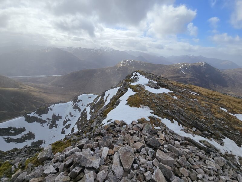 A crest of broken pale stone with old snow, falling steeply on the left, and ranges under cloud beyond.