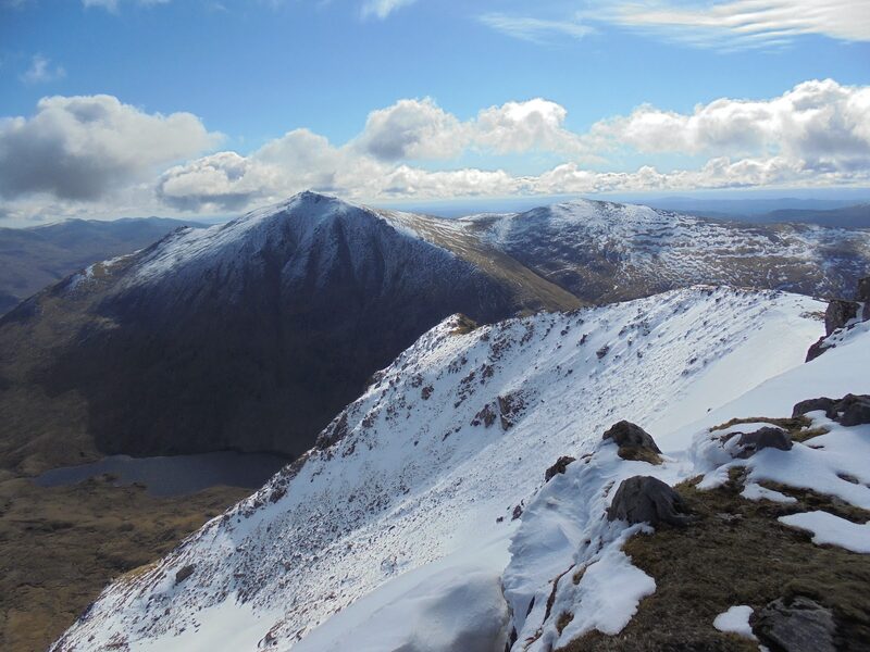 Sgurr na Lapaich under a dusting of snow, seen across a deep gap from a snow-covered ridge, with a lochan below.