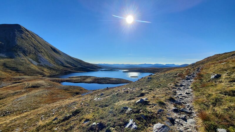 A stony path dropping over tawny autumn grass to the lochans under the hill, its scree flank on the left and far blue ranges beyond, into the sun.