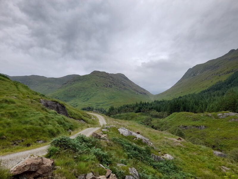 A gravel track winding up a green glen towards a rounded hill under grey cloud.