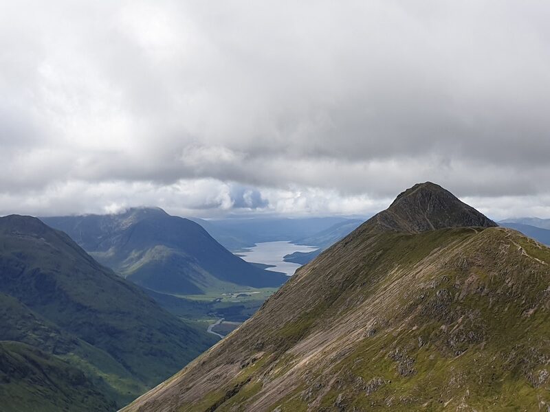 A pointed summit on a grassy ridge, with a sea loch far below in the glen beyond and heavy cloud overhead.