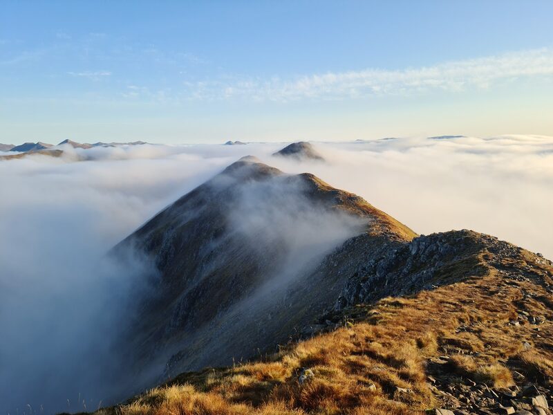 A narrow ridge running out above a sea of cloud to a further top in early light.