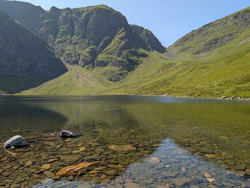 The cliffs of Coire Ardair in sun above their still lochan, stones showing through the clear water in front and the notch of the Window on the skyline.