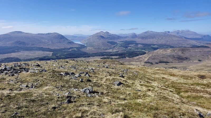 From a grassy, stony shoulder across forestry to a loch in a trench between hills, a twin-topped hill to its right and snow-flecked hills far off.