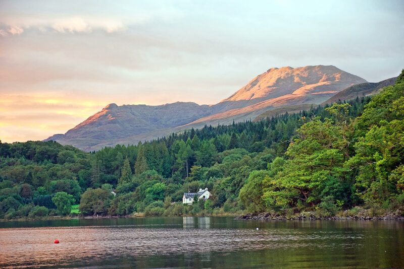 The hill lit at sunset above a wooded loch shore and a white house.