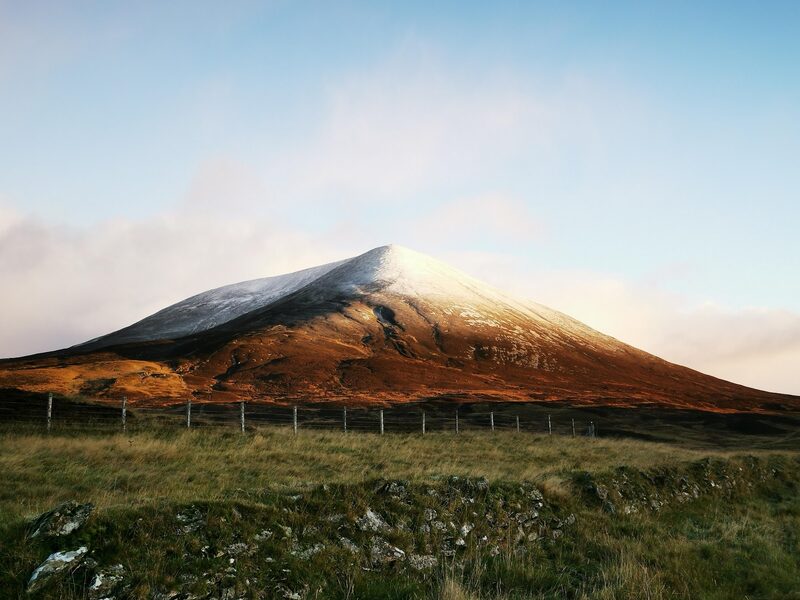 Càrn Liath's cone under a cap of snow, lit russet by a low sun, above a fence line and rough grass.