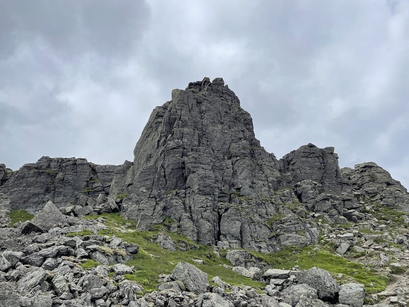 A prow of dark rock standing over boulders and grass high on Beinn Narnain, under grey cloud.