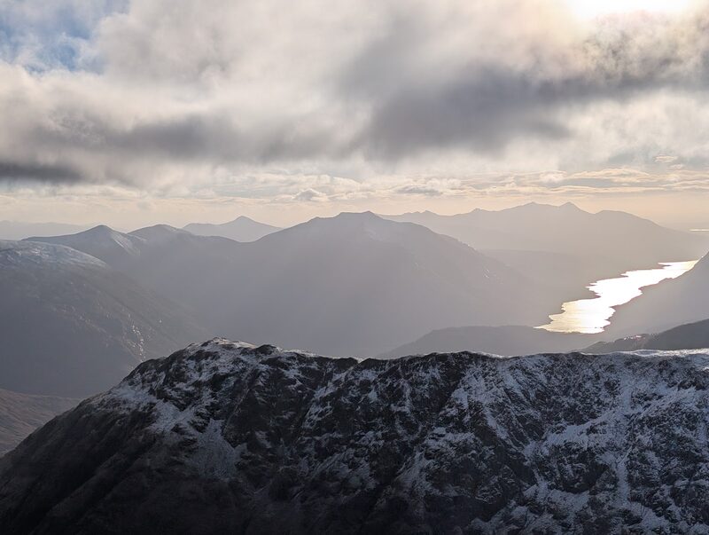 A snow-dusted ridge in the foreground and a sea loch shining far below, hills in silhouette under winter cloud.