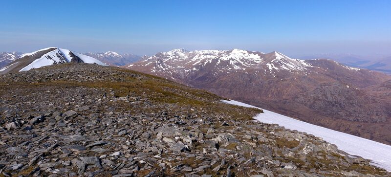 Stony summit ground with a patch of old snow, and snow-streaked ranges beyond under a clear sky