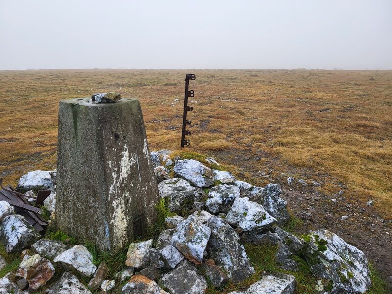 The trig pillar in its pile of white quartz, an old iron fence post beside it and flat moor fading into mist.