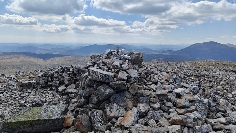 The summit cairn on stony ground, with hills beyond under broken cloud.
