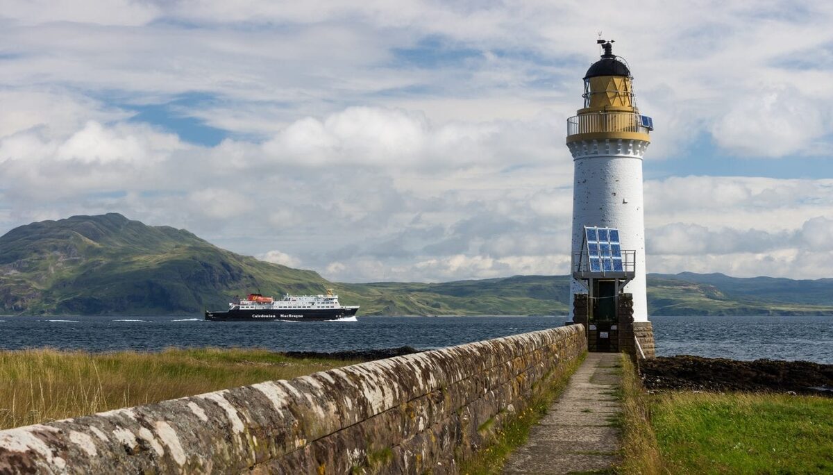 A Caledonian MacBrayne ferry passes a lighthouse along a stone harbour wall