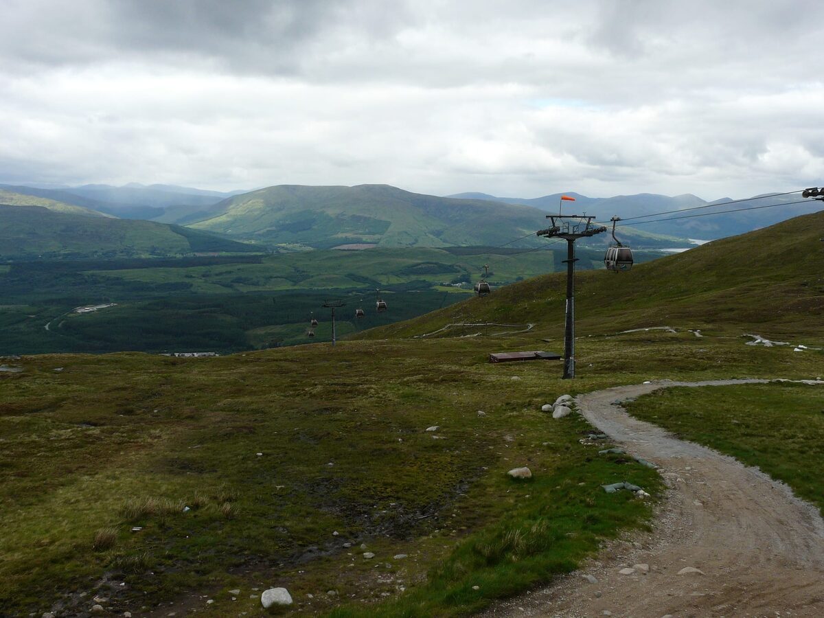 Chairlift pylons above a gravel path overlooking layered glen and mountains
