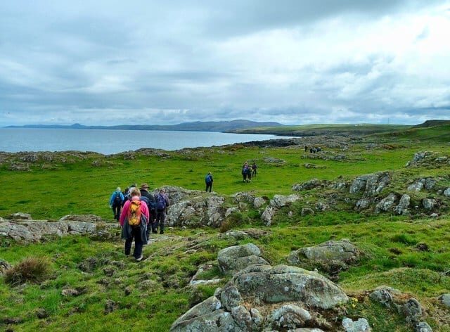 Walkers cross rocky coastal grassland towards a distant headland under grey skies