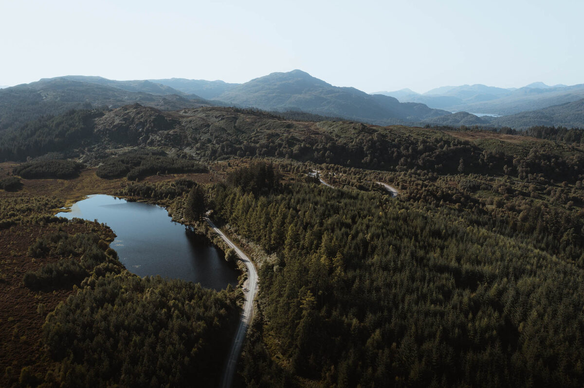 The forest drive winding past a loch through the woods, from above.