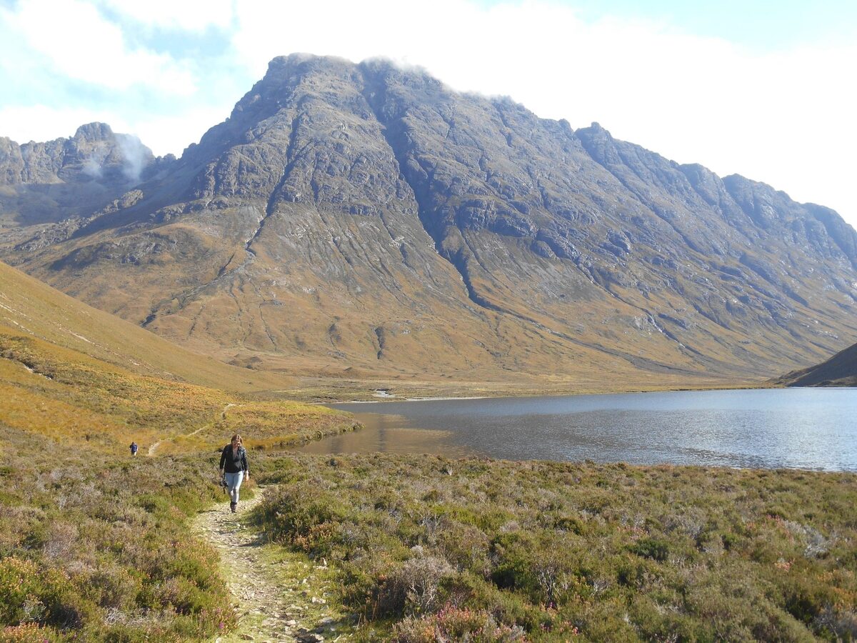 Walker on a path beside a loch below a steep mountain