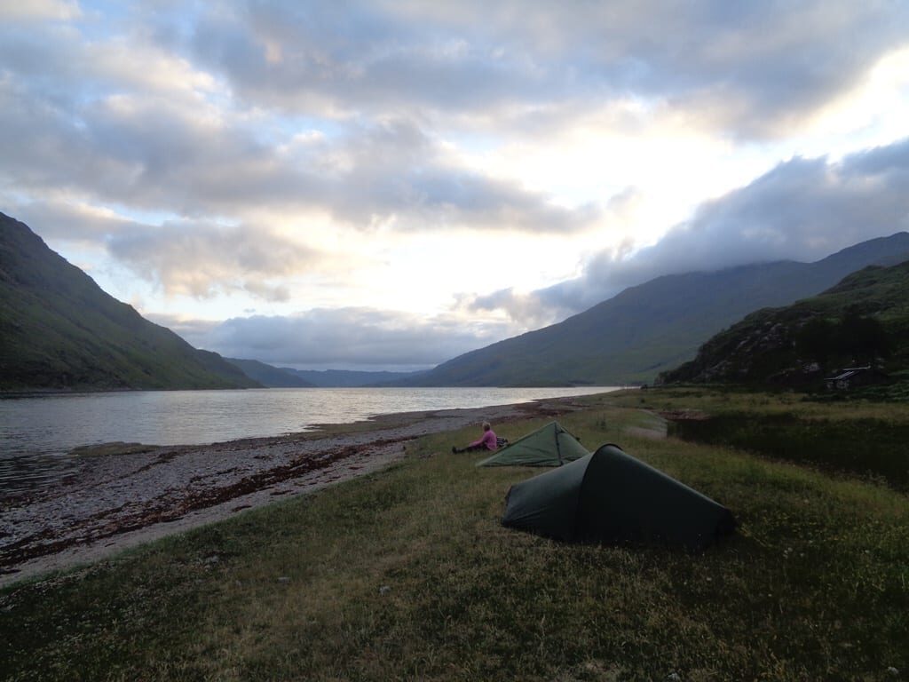 Green tent pitched by the shore of a loch flanked by hills
