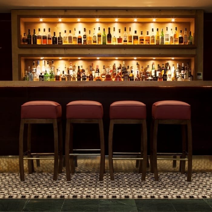 Bar stools face shelves of spirits lit above a patterned tiled floor
