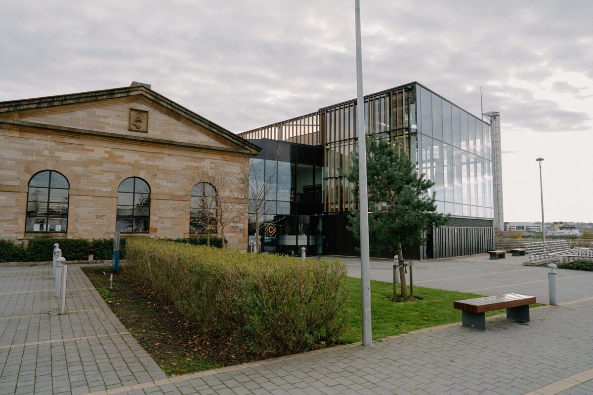 Sandstone building beside a modern glass extension with a wooden bench nearby