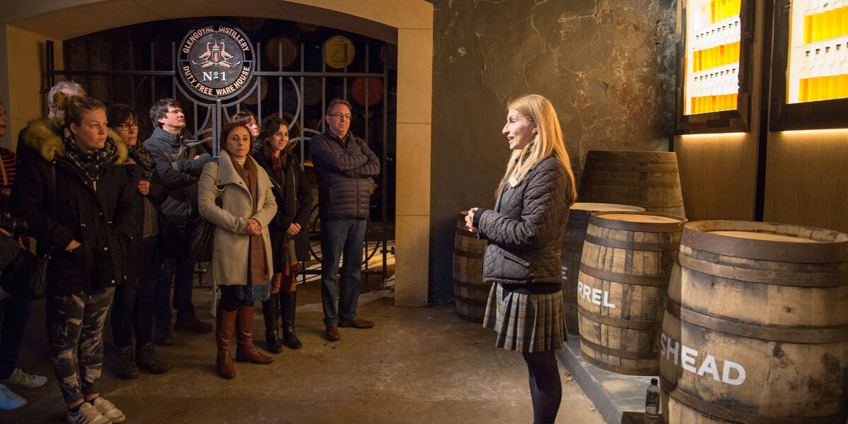 Guide addresses visitors beside whisky barrels marked "HEAD" in a distillery hall