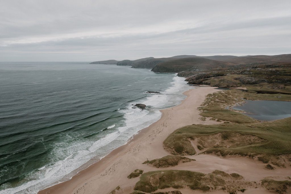 Aerial view of a curving sandy beach with dunes and coastal hills