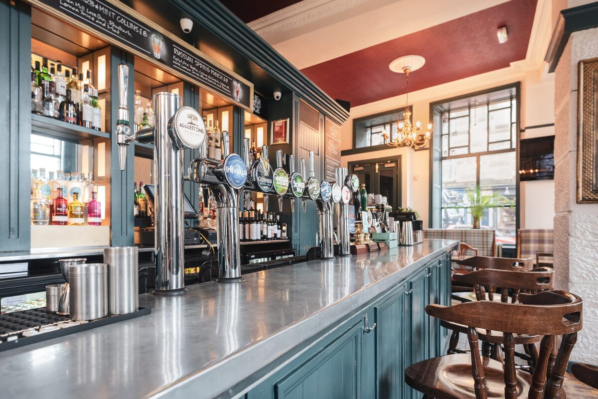 Rows of beer taps line a pub bar with dark green cabinetry and wooden stools