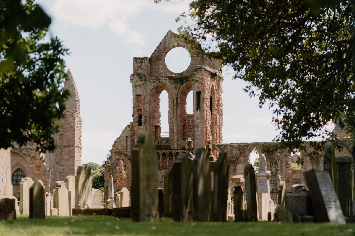Ruined red sandstone abbey with round window, gravestones in the foreground