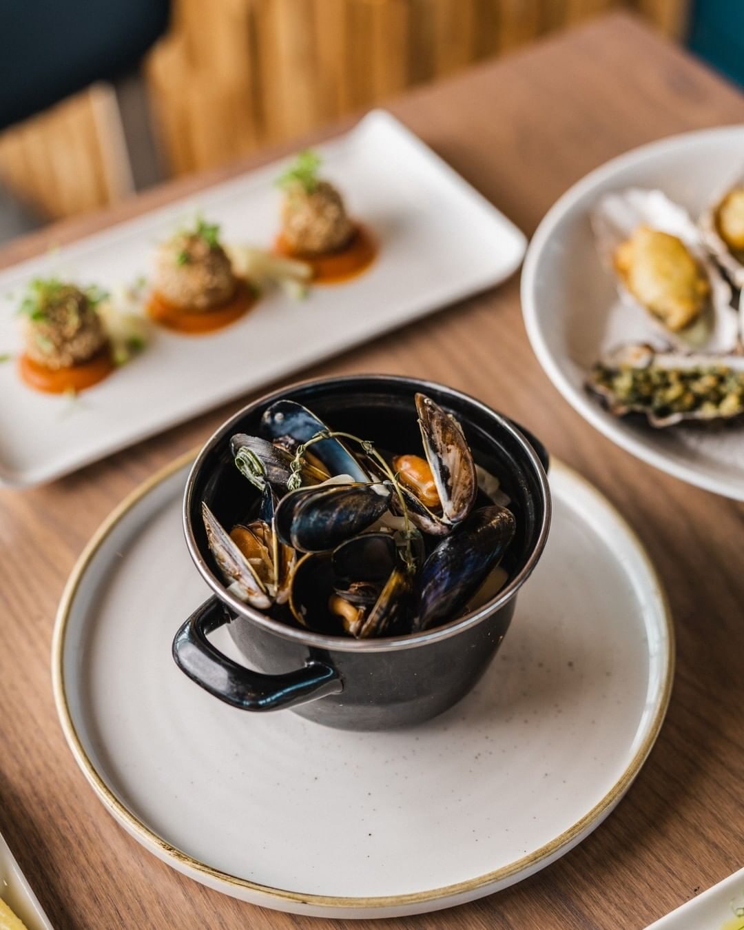 Mussels in a black pot beside breaded croquettes and dressed oysters on a wooden table