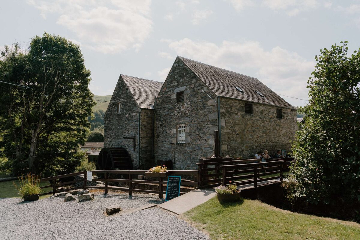 Stone mill building with waterwheel beside a wooden footbridge and river