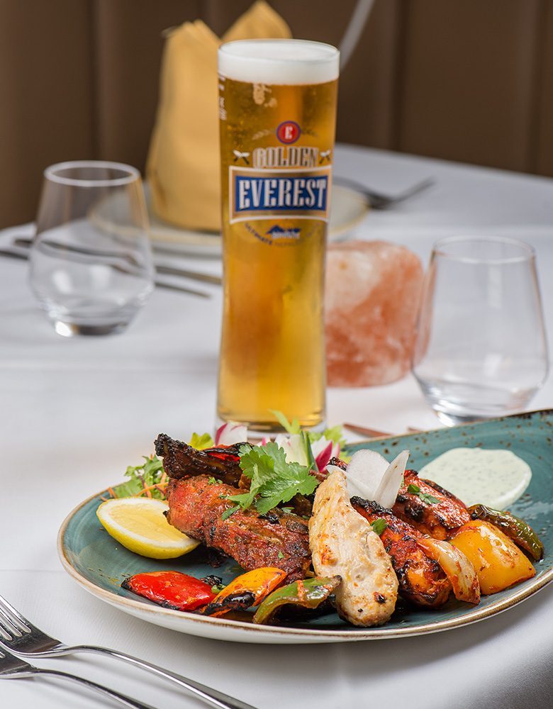Golden Everest beer beside a plate of grilled tandoori chicken and peppers