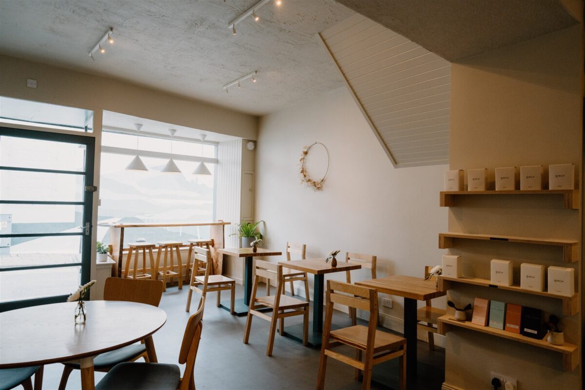 Wooden tables and chairs in a bright café, shelves stacked with boxed goods