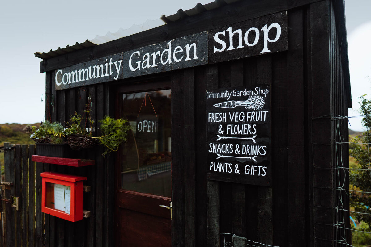 The black timber Community Garden Shop beside the road, its painted sign listing fresh veg, fruit and flowers.