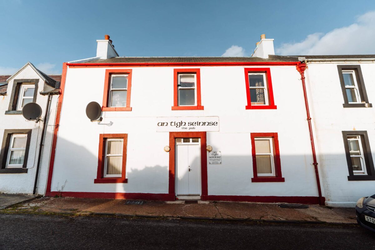 The whitewashed front of An Tigh Seinnse in Portnahaven, its windows and door picked out in red.