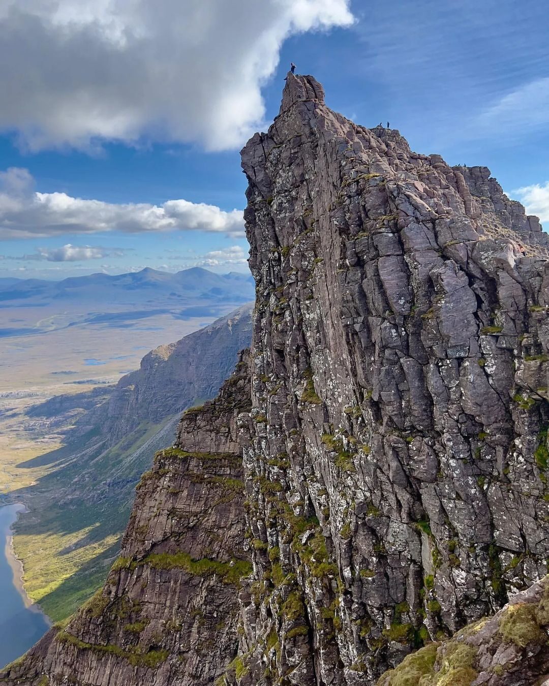 A climber stands atop a jagged rock pinnacle above a glen and loch