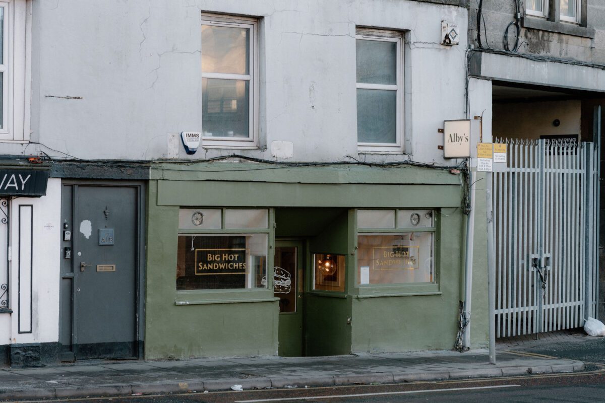 Green-fronted shopfront reading "Big Hot Sandwiches" beside a weathered white tenement building