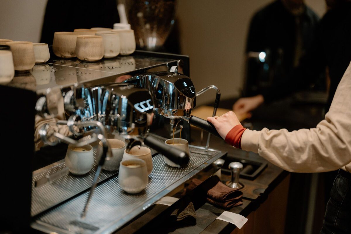 Barista pulling espresso shots on a chrome machine with stacked cups nearby