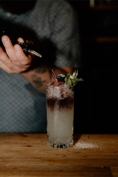 Bartender pours a dark liqueur over crushed ice onto a cocktail garnished with mint and blackberry