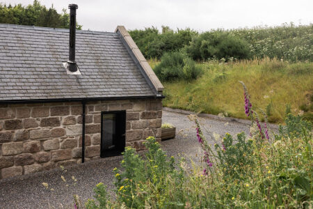 A stone cottage with a slated roof and black stove pipe sits beside a gravel drive, foxgloves and wildflowers lining the path.