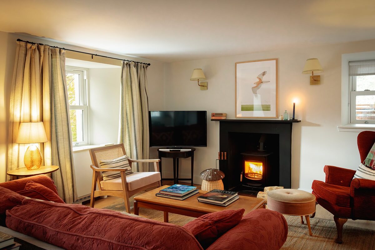 A red sofa and armchairs surround a low table in a sitting room lit by lamps, a wood burning stove alight in the fireplace opposite.