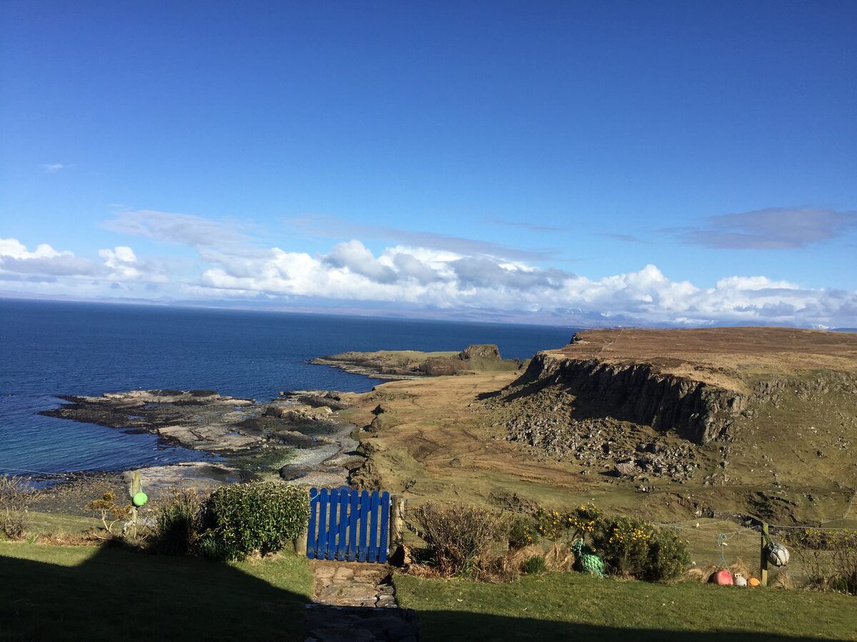 A weathered blue gate looks out over a rocky coastline and calm sea under a clear sky.