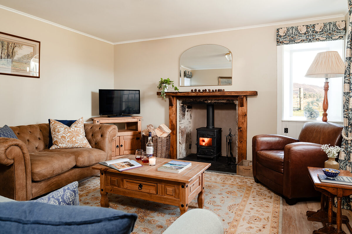 A wood burning stove burns in a stone fireplace beneath an arched mirror, a leather sofa and armchair arranged around a wooden coffee table.