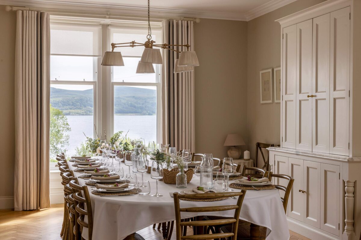 A long dining table set for a group stands beneath brass pendant lights, a loch and hills visible through the window and a panelled cabinet against the wall.