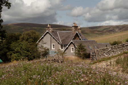 A grey stone farmhouse with several chimneys stands against rolling hills, a wildflower meadow in the foreground.
