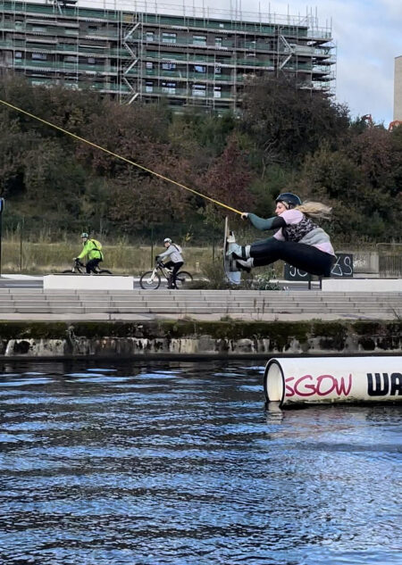 A wakeboarder in the air behind the cable, riders waiting on the pontoon and a tower block behind.
