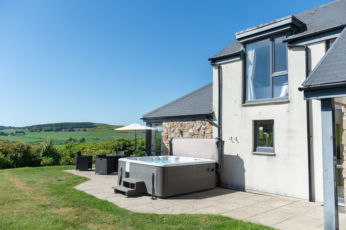 A hot tub with its cover open sits on a patio beside a stone and render house, rolling countryside visible in the distance.