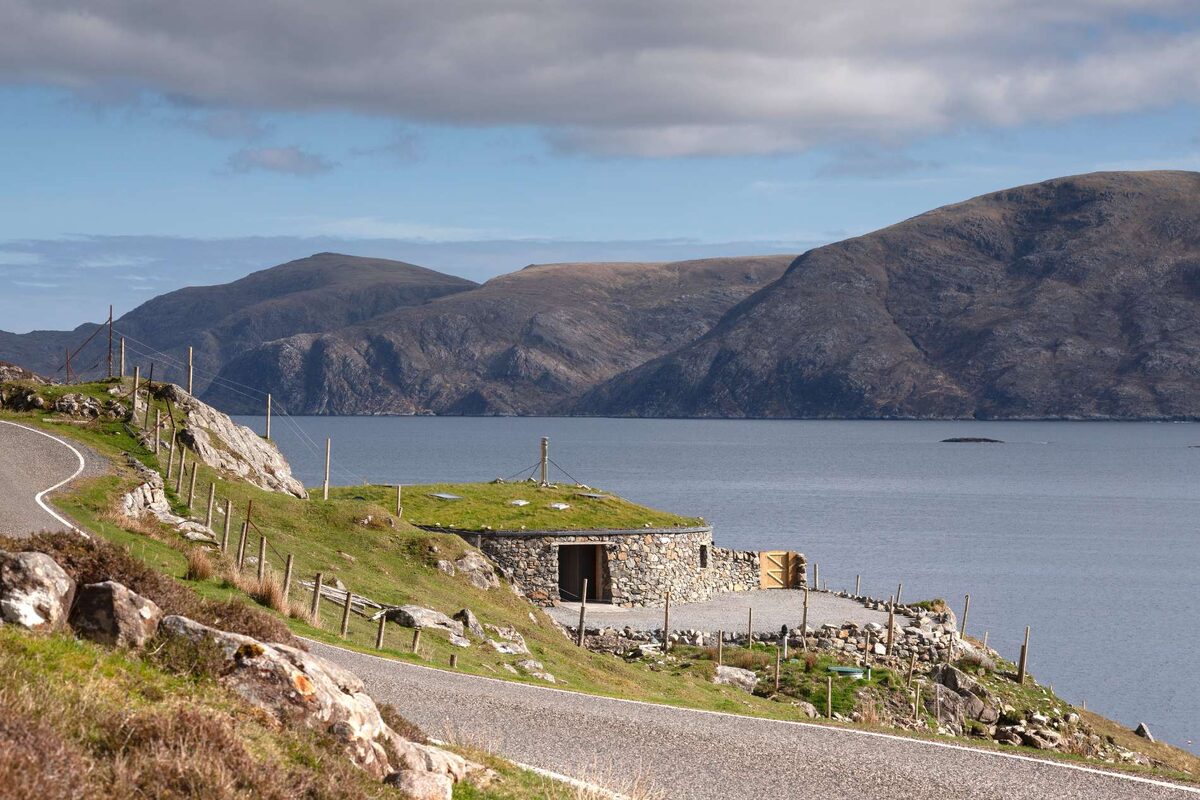 A stone bothy with a turf roof sits on a headland overlooking a sea loch framed by hills.