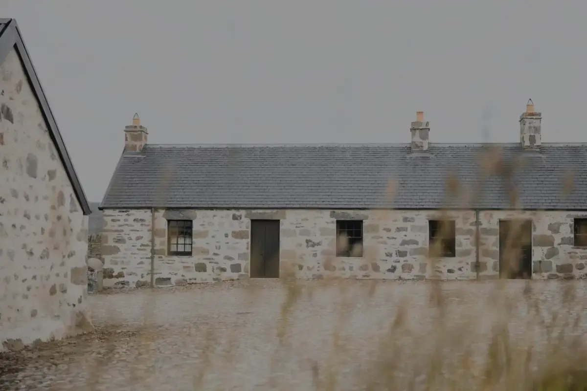 A row of stone cottages with a slated roof and chimneys stands behind a gravel yard, seen through tall grass.