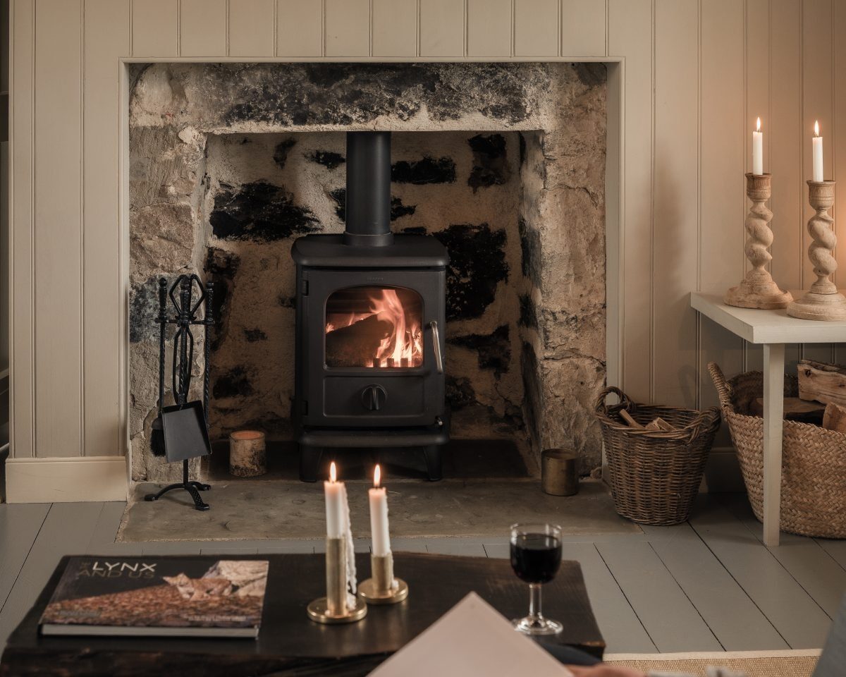 A wood burning stove is alight in a stone fireplace, lit candles and a glass of wine on the table in front beside a copy of "Lynx" magazine.