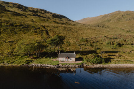 An aerial view shows a white cottage on a wooded loch shore, hills rising steeply behind it.