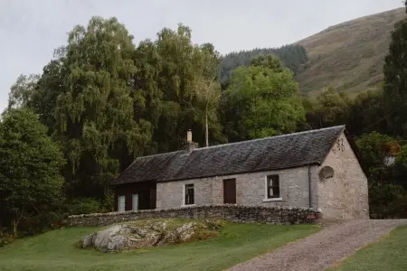 A stone cottage with a slate roof and chimney stands among birch and pine trees on a wooded hillside.